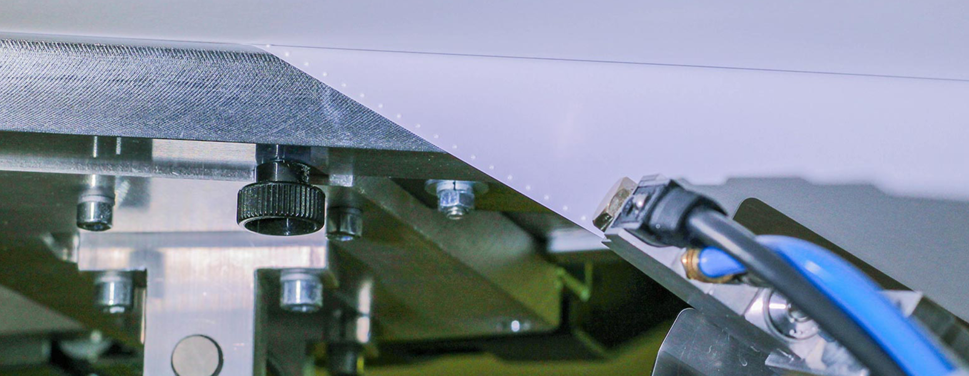 ALFAST® 14 Das Bild zeigt einen Ausschnitt einer industriellen Druckverarbeitungsmaschine, die für den Auftrag von Klebstoff verwendet wird. Im Fokus steht der präzise Mechanismus der Maschine, der aus mehreren Metallteilen, Schrauben und Justierschrauben besteht. Ein blaues Kabel und andere mechanische Komponenten sind ebenfalls sichtbar. Die Oberfläche, auf die der Klebstoff aufgetragen wird, ist glatt und weiß, mit kleinen punktierten Markierungen entlang einer Kante, die auf die Präzision des Klebevorgangs hinweisen. Das Bild vermittelt ein Gefühl von technischer Raffinesse und Präzision in der Druckverarbeitung.